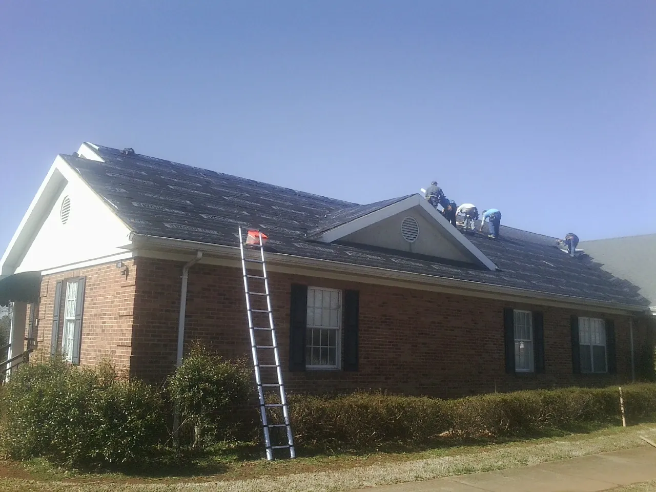 Crew tearing shingles off a brick ranch roof with a ladder set — Metal Roof Repair work by our crew in Cave Spring